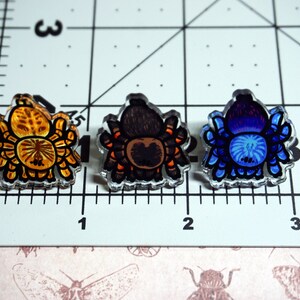 May include: Three colorful tarantula pins. The first pin is orange and yellow, the second is brown and orange, and the third is blue and purple.