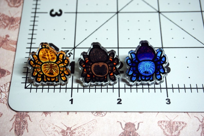 May include: Three colorful tarantula pins. The first pin is orange and yellow, the second is brown and orange, and the third is blue and purple.