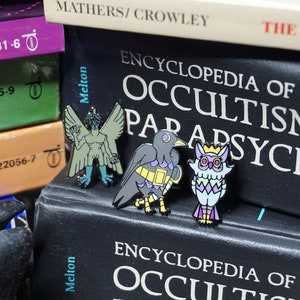 May include: Three enamel pins featuring a green winged demon, a black bird with yellow and purple accents, and a purple and green owl with a gold crown. The pins are displayed on a black book with the text "ENCYCLOPEDIA OF OCCULTISM & PARAPSYCHOL" on the cover.
