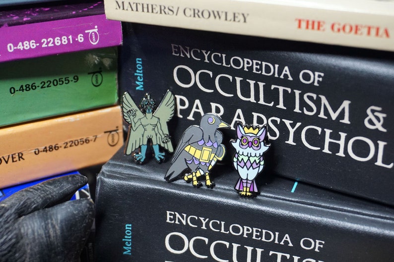 May include: Three enamel pins featuring a green winged demon, a black bird with yellow and purple accents, and a purple and green owl with a gold crown. The pins are displayed on a black book with the text "ENCYCLOPEDIA OF OCCULTISM & PARAPSYCHOL" on the cover.