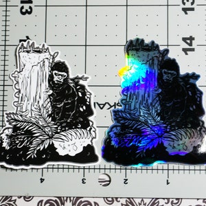 May include: Two black and white stickers of a gorilla standing in front of a tree. One sticker is a standard black and white print, and the other is a holographic sticker with a rainbow sheen.