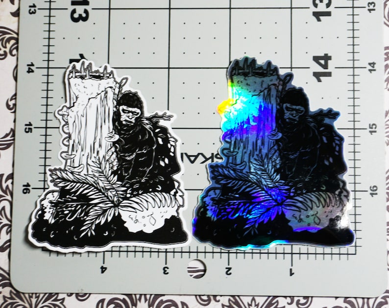 May include: Two black and white stickers of a gorilla standing in front of a tree. One sticker is a standard black and white print, and the other is a holographic sticker with a rainbow sheen.