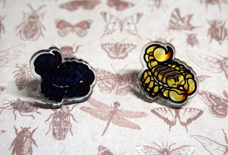 May include: Two clear acrylic pins featuring cartoon scorpions. One scorpion is blue and the other is yellow.