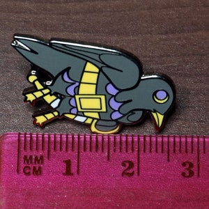 May include: A black and purple enamel pin of a crow with yellow accents. The crow is wearing a yellow and purple band around its body. The pin is about 2 inches long.