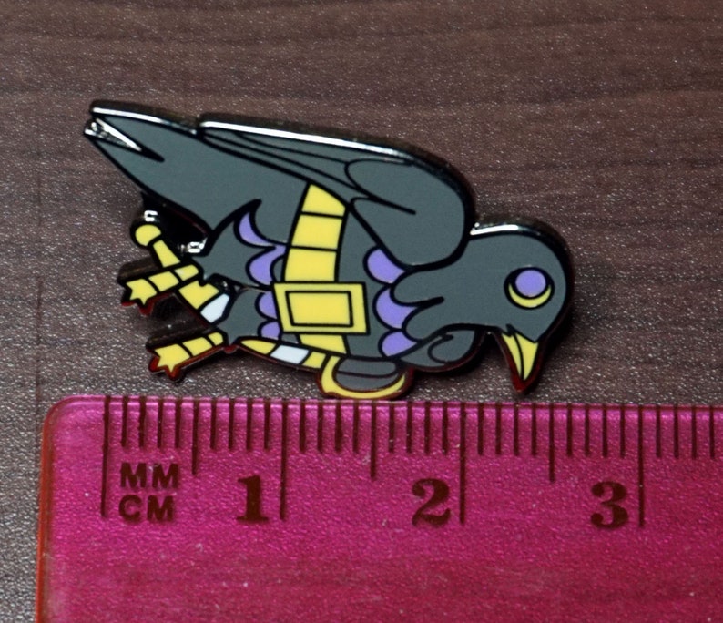 May include: A black and purple enamel pin of a crow with yellow accents. The crow is wearing a yellow and purple band around its body. The pin is about 2 inches long.