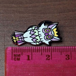 May include: A purple, green, and yellow enamel pin of an owl wearing a crown. The owl has large, round eyes with yellow pupils. The pin is approximately 1 inch in size.