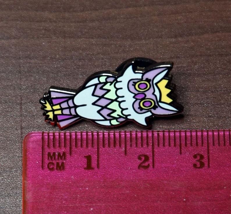 May include: A purple, green, and yellow enamel pin of an owl wearing a crown. The owl has large, round eyes with yellow pupils. The pin is approximately 1 inch in size.