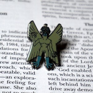 May include: A green and blue enamel pin depicting a winged humanoid creature with a grotesque face. The creature has a human-like body with wings and clawed hands.