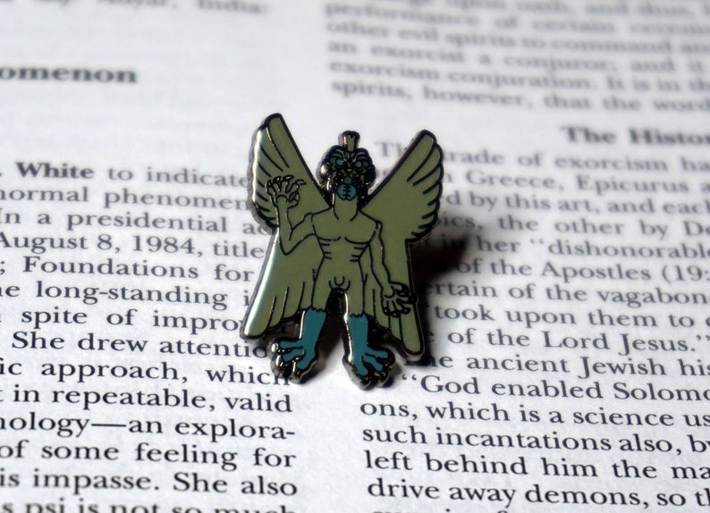 May include: A green and blue enamel pin depicting a winged humanoid creature with a grotesque face. The creature has a human-like body with wings and clawed hands.