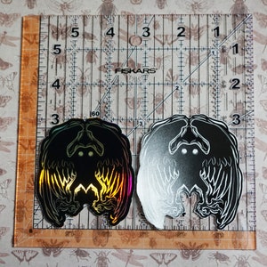 May include: Two black and white stickers with a holographic finish. The stickers feature a creature with wings and glowing eyes. The stickers are on a ruler with inch markings.