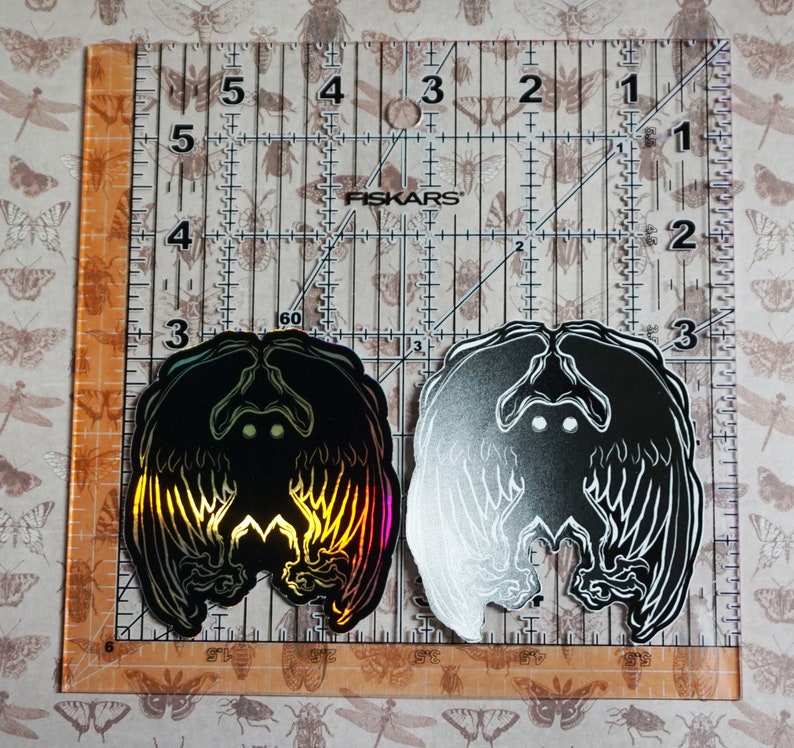May include: Two black and white stickers with a holographic finish. The stickers feature a creature with wings and glowing eyes. The stickers are on a ruler with inch markings.