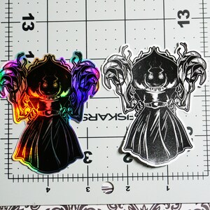 May include: Two black and white stickers of a cartoon character with a large head and a long black robe. The character has a large, round head with two eyes and a wide mouth. The character is wearing a long black robe with a hood. The sticker on the left is iridescent and has a rainbow sheen. The sticker on the right is black and white.