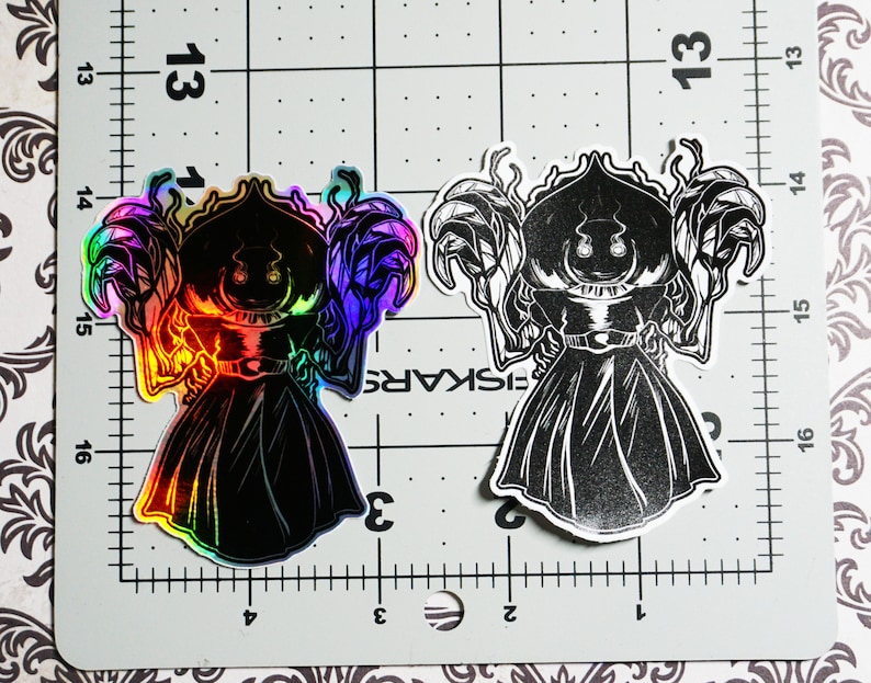 May include: Two black and white stickers of a cartoon character with a large head and a long black robe. The character has a large, round head with two eyes and a wide mouth. The character is wearing a long black robe with a hood. The sticker on the left is iridescent and has a rainbow sheen. The sticker on the right is black and white.