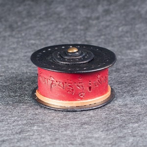 May include: A cylindrical prayer wheel with a black top and base, and a vibrant red center. The red section features embossed text and is accented with a gold band. A small brass-colored stud is at the center of the top.