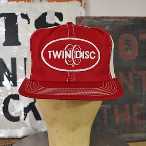 May include: Red and white trucker hat with the text "TWIN DISC" in a white oval. The hat has a red front panel and brim, with a white mesh back. The hat is displayed on a mannequin head.