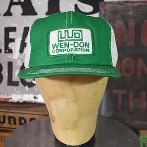 Op de afbeelding: Groene en witte truckerpet met de tekst "WEN-DON CORPORATION" op een witte patch. De pet heeft een groen voorpaneel en klep, met een witte mesh achterkant. De pet wordt getoond op een mannequinhoofd.