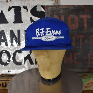 Pode incluir: Boné de beisebol azul com o texto "B.F. Evans Ford Kentucky" em branco e um logotipo oval branco. O boné é exibido em uma cabeça de manequim. O fundo inclui sinais vintage.