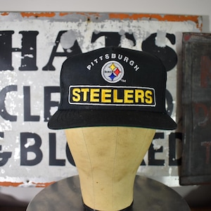 May include: A black baseball cap featuring the Pittsburgh Steelers logo embroidered on the front. The word Pittsburgh is arched above the circular team logo, and the word Steelers is displayed in bold yellow letters on a black rectangular patch.