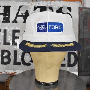May include: A white baseball cap featuring a blue rectangular patch with the Ford logo and the word FORD in white text. The cap has a dark blue brim adorned with gold-colored embroidered laurel leaf detailing along the front edge of the brim.