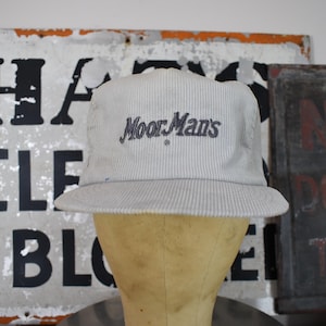 May include: A light gray corduroy baseball cap featuring the text MoorMan's embroidered in dark blue thread on the front panel. The hat has a structured crown, a curved brim, and a textured fabric surface with vertical ridges throughout the design.