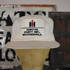Puede incluir: Gorra de béisbol color crema con visera curva. La gorra presenta un parche rectangular con el texto "HAMILTON COUNTY IMPL. MCLEANSBORO, IL." y un logotipo. El sombrero se muestra en una cabeza de maniquí.