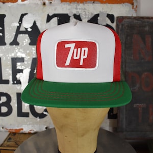 May include: A white, red, and green trucker hat featuring a rectangular patch on the front panel with the text 7up in white letters on a red background. The cap has a solid white front panel, red side panels, and a green brim with stitching.