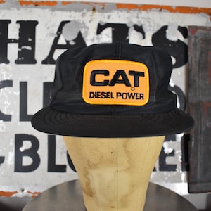 May include: A black baseball cap featuring a rectangular patch on the front. The patch has a yellow background with a black border and displays the text CAT in large letters above the words DIESEL POWER in smaller black capital letters.