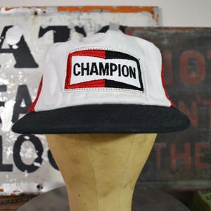 May include: A Champion baseball cap in white, red, and black. The cap has the word "CHAMPION" in black on a white patch, framed by red and black. The brim is black. The cap is displayed on a mannequin head.