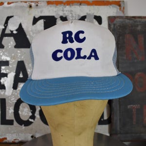 May include: A vintage RC Cola trucker hat with a white front panel and a blue brim. The hat features the text "RC COLA" in blue block letters. The hat has a mesh back and is displayed on a mannequin head.