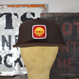 Puede incluir: Gorra de béisbol marrón con visera plana y parche con el logotipo de Shell. El parche presenta un diseño de concha amarilla sobre fondo rojo con borde blanco. La gorra se muestra sobre una cabeza de maniquí.