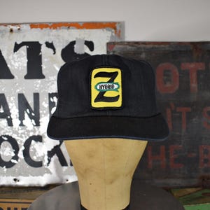 May include: Black cap with a yellow and green patch that reads "HYBRID" and features a stylized "Z" design. The cap has a curved brim and a blue trim. The cap is displayed on a mannequin head.