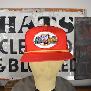 May include: A bright red trucker hat featuring a mesh back and a yellow cord trim along the base of the crown. The front displays a white oval patch with a Union Pacific logo, depicting a black steam locomotive and a yellow diesel train engine.