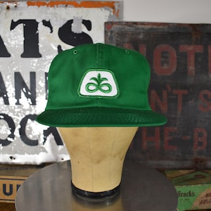 Può includere: Cappellino da baseball verde con una toppa con logo bianco e verde. Il cappellino è esposto su una testa di manichino. Lo sfondo include insegne vintage e una scatola di mentine.