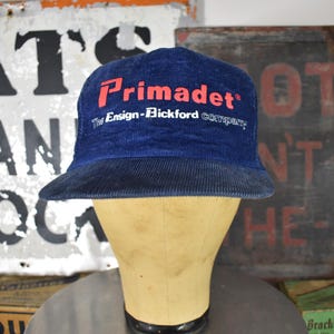 May include: A navy blue corduroy cap with the word "Primadet" in red lettering. The text "The Ensign-Bickford Company" is below the main text. The hat is displayed on a mannequin head.