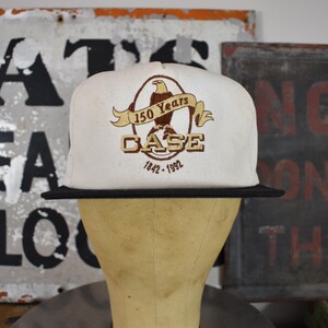 May include: A white baseball cap with a black peak, displaying an embroidered logo with an eagle, banner, and the text "150 Years CASE 1842-1992". The cap is shown on a mannequin head against a backdrop of vintage signs.