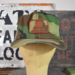 May include: Camouflage baseball cap with a brown peak and a red patch that reads "BUNGE". The cap is displayed on a mannequin head. The background includes vintage signs with faded lettering.