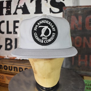 May include: A light grey trucker cap with a white mesh back and a curved peak. The front has a black and white circular patch reading "70th Anniversary Gleaner Combine 1993". The cap is displayed on a mannequin head.