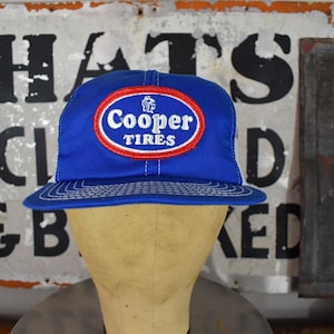 May include: A blue Cooper Tires baseball cap with a white stitched brim and a red and white logo. The cap is displayed on a mannequin head. Vintage sign in the background.