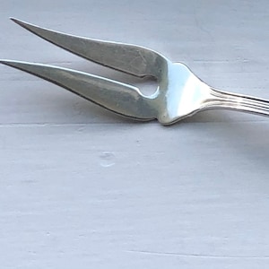 May include: A silver two-pronged fork with a decorative handle. The fork is designed for serving food.