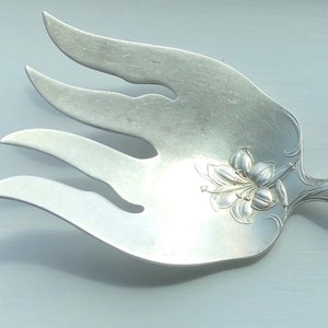 Puede incluir: Un tenedor de servir de plata con un diseño floral decorativo en el mango. El tenedor tiene tres dientes y una cabeza ancha y plana.