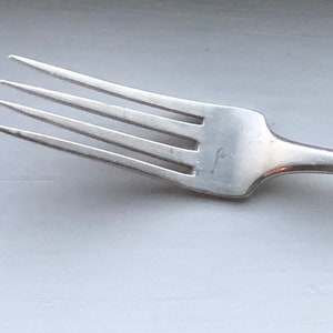 May include: A silver fork with a simple design. The fork has four tines and a rounded handle.