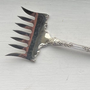 May include: Silver fork with a decorative handle featuring a floral design. The fork has a unique design with multiple tines.