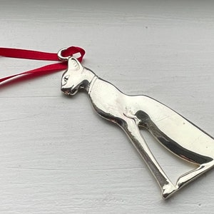 May include: Silver cat ornament with a red ribbon. The cat is sitting with its tail curled around its legs.
