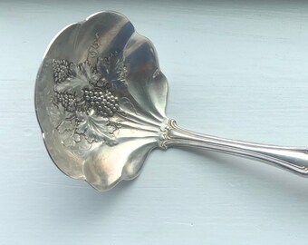 May include: A silver serving spoon with an ornate design featuring grapes and leaves. The spoon has a wide, rounded bowl and a long, curved handle.