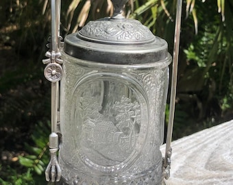 May include: A silver and glass antique sugar dispenser with an ornate design. The dispenser has a glass jar with a silver lid and a silver handle. The handle is shaped like an arch and has a decorative design.