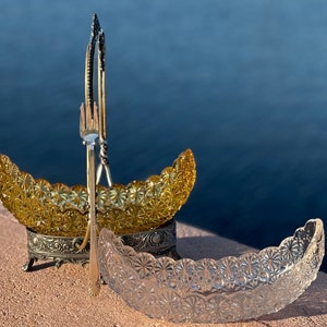 May include: A silver serving set with two crescent-shaped glass bowls. The larger bowl is amber glass with a floral pattern and the smaller bowl is clear glass with a similar pattern. The set is on a wooden surface.