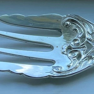May include: Silver serving fork with three tines and ornate, scrolling floral designs. The fork features a handle with detailed embellishments and a decorative, wing-like element on each side of the tines. A classic piece for serving.