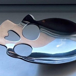 May include: A silver spoon with a skull-shaped design and three holes in the bowl. The spoon has a long, curved handle.