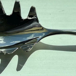 Op de afbeelding: Een zilveren saladelepel met een decoratief handvat en een uniek, meerpuntig ontwerp.