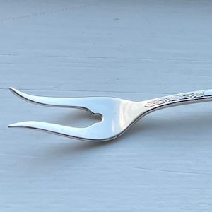 May include: A silver fork with a decorative handle and three prongs. The fork is designed for serving food.